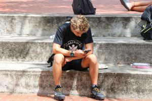 A student dressed in shorts and a t-shirt sits on concrete steps looking at his phone.